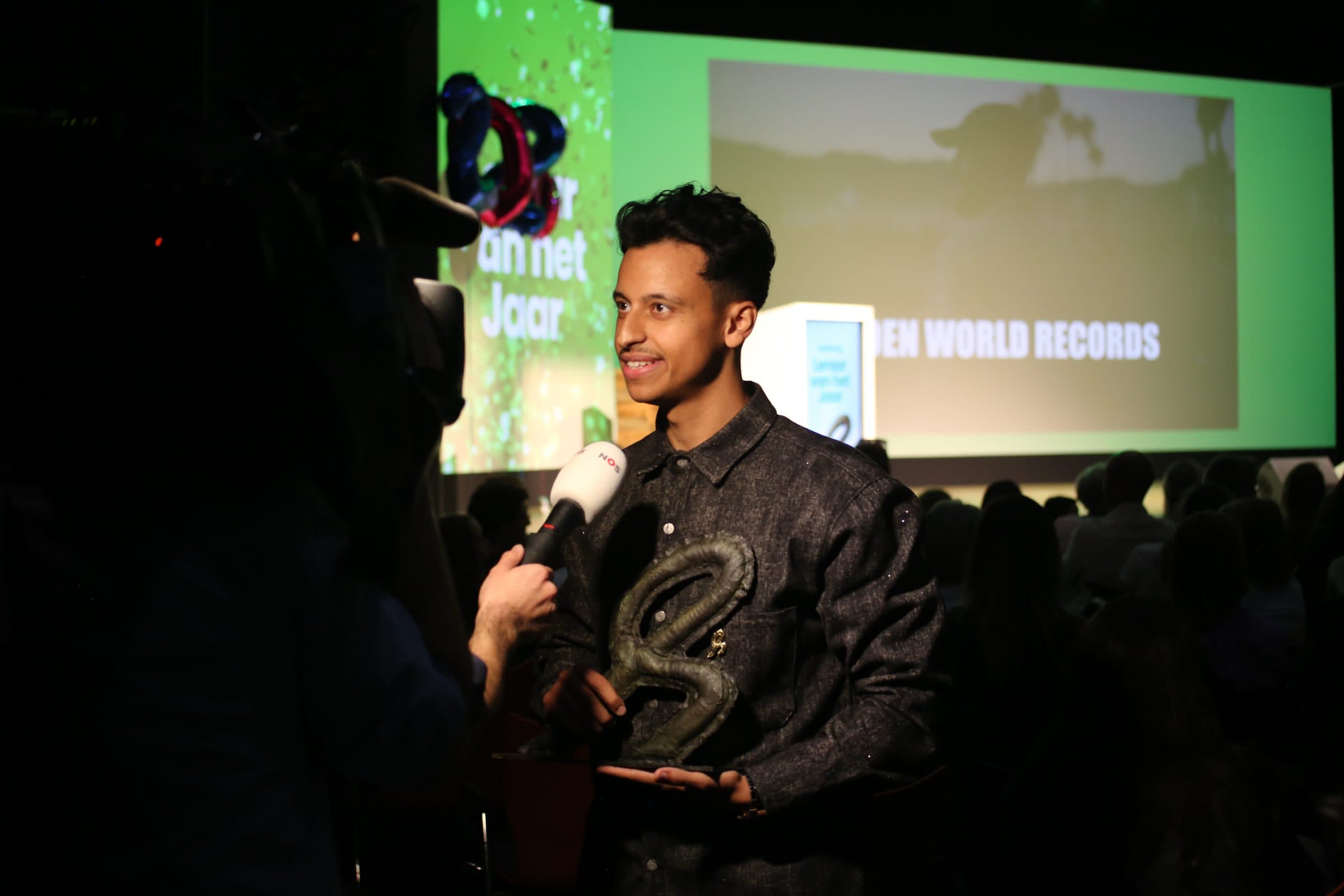 Bilal op een donker podium met de trofee, terwijl hij via een microfoon wordt geïnterviewd