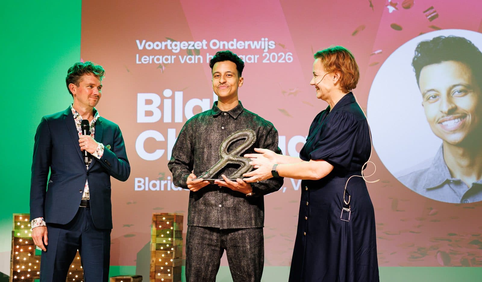 Bilal staat met zijn trofee tussen de presentatoren voor het scherm 'Voortgezet Onderwijs, Leraar van het Jaar 2026'