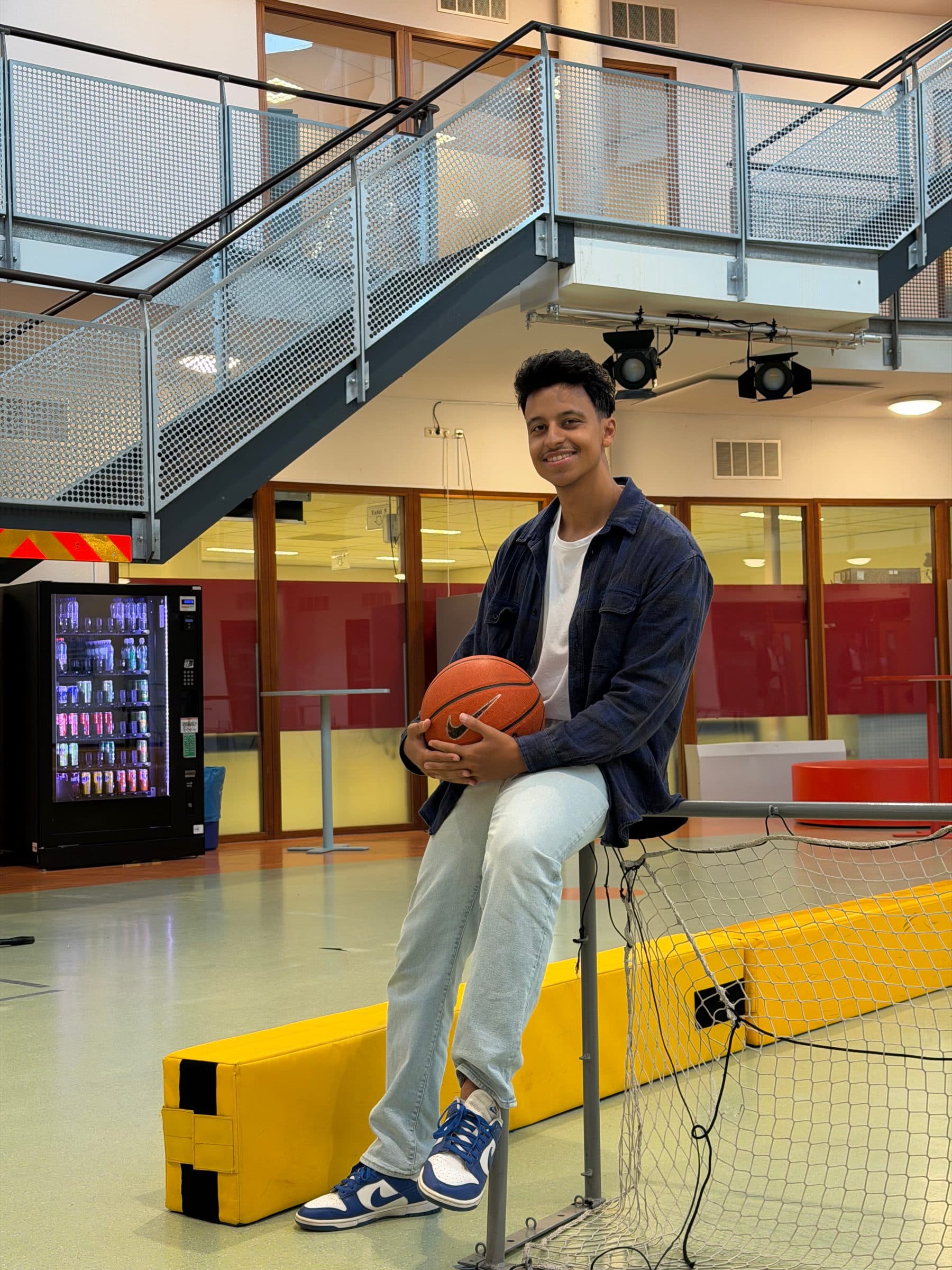Bilal op een kruk met een basketbal in de aula van school